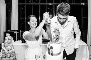 Un couple de mariés découpe avec enthousiasme un gâteau décoratif lors d'une réception de mariage. Le gâteau est composé de plusieurs étages surmontés de figurines. Un autre dessert est visible sur la table à côté.