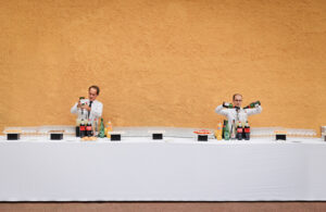 Deux serveurs en uniformes blancs servent des boissons derrière une longue table recouverte d'une nappe blanche. La table est dressée avec des verres, des assiettes et des bouteilles. L'arrière-plan présente un mur jaune texturé et chaleureux.
