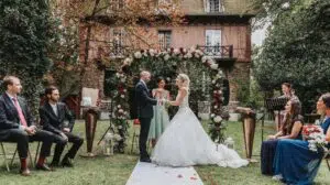 Une mariée et un marié se tiennent sous une arche florale échangeant leurs vœux lors d'une cérémonie laïque de mariage, entourés de leurs témoins de chaque côté, avec un manoir rustique en arrière-plan.