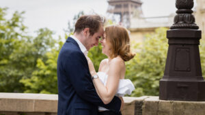 Un couple se tient debout, l'un près de l'autre, souriant et se touchant le front, vêtu d'une tenue de soirée. Ils paraissent heureux et affectueux, avec de la verdure et des détails architecturaux en arrière-plan.