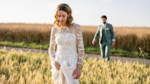 Une mariée vêtue d'une robe de mariée en dentelle blanche marche dans un champ ensoleillé en tenant sa jupe, tandis qu'un marié en costume vert la suit à l'arrière-plan.
