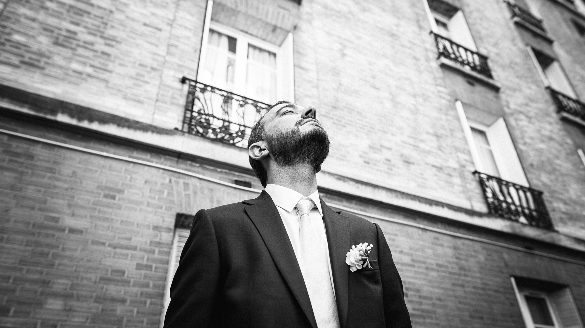 Le premier regard, le marié en costume avec une boutonnière regarde vers le haut, debout devant un bâtiment aux murs de briques et aux balcons de fer. La photo est en noir et blanc, ce qui crée une atmosphère dramatique et formelle.