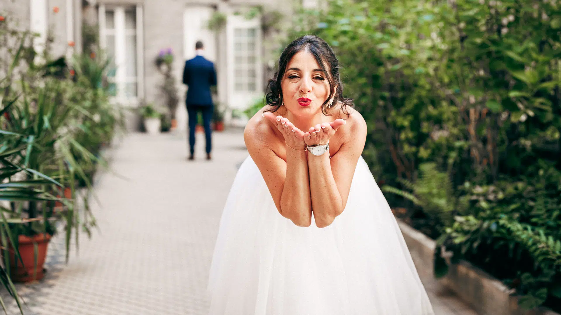 Une mariée en robe blanche envoie un baiser à l'appareil photo dans un jardin extérieur, tandis qu'un homme en costume se tient à l'arrière-plan, de face.