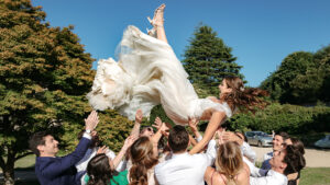 Un groupe de personnes en tenue de soirée lance joyeusement une mariée souriante vêtue d'une robe blanche dans les airs, par une journée ensoleillée, avec des arbres verts et un ciel bleu en arrière-plan.