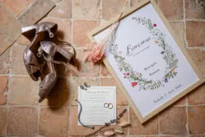 Flat lay mariage élégant avec chaussures à talons, affiche d’accueil florale encadrée, invitation délicate, alliances et bijoux sur fond rustique.