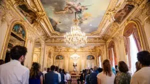 Un groupe de personnes est assis face à un couple, au fond d'une salle somptueuse aux moulures dorées, ornée d'un lustre et d'un plafond peint, lors d'une cérémonie civile de mariage à la mairie du 8e arrondissement de Paris.