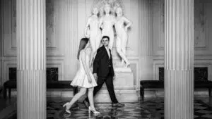 Une mariée vêtue d'une robe légère et un marié en costume marchent main dans la main devant des statues classiques dans une élégante salle ornée de colonnes de la mairie du 8e arrondissement de Paris.