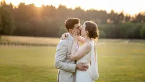 Un couple en tenue de mariage s'embrasse dans un champ baigné de soleil, avec des arbres en arrière-plan. La mariée porte une robe blanche et le marié un costume clair. La scène dégage une atmosphère romantique et sereine.