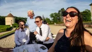 Quatre personnes sont assises à une table en terrasse ; elles rient et s'amusent sous le soleil. Trois hommes portent des chemises et des lunettes de soleil, et une femme en robe bleue porte également des lunettes de soleil. Des verres vides sont posés sur la table. La scène se passe au château de Breteuil.