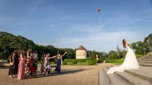 Une mariée vêtue d'une robe blanche lance son bouquet haut dans les airs tandis qu'un groupe de femmes s'empresse de l'attraper sur une allée pavée, dans le cadre pittoresque du jardin du château de Breteuil.