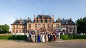 Un grand groupe de personnes en tenue de cérémonie pose pour une photo devant le majestueux château de Breteuil dans les Yvelines aux briques orange et au toit d'ardoise, par une journée ensoleillée.