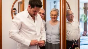 Le marié boutonne sa chemise tandis que ses parents, souriant, l'observe dans une pièce baignée d'une lumière chaleureuse, décorée de miroirs et de cadres en bois. On aperçoit un autre homme dans l'embrasure de la porte, à l'arrière-plan.