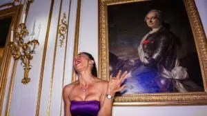 Une femme vêtue d'une robe violette sans bretelles rit joyeusement à côté d'un grand portrait richement orné représentant un homme en armure ; tous deux se trouvent au château de Breteuil dans les Yvelines, dans un décor raffiné rehaussé de dorures.