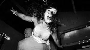 Une femme aux cheveux en bataille, arborant un large sourire, est soulevée dans les airs sur dancefloor, les bras tendus. La scène est en noir et blanc et rend compte d'un mouvement énergique et joyeux.