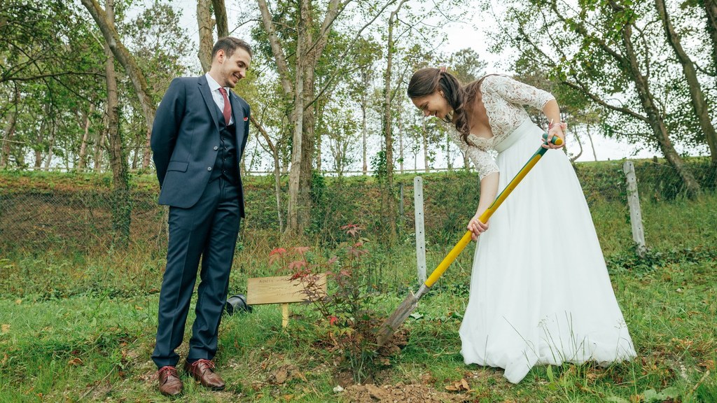 Une mariée en robe blanche plante un petit arbre à l'aide d'une pelle, tandis qu'un marié en costume la regarde en souriant, à l'extérieur, lors du rituel de l'arbre planté.