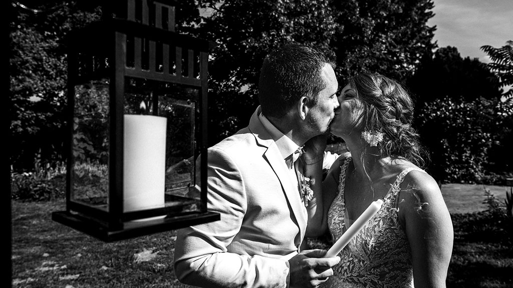 Un couple en tenue de mariage s'embrasse en plein air, une bougie à la main pour le rituel des bougies. Une lanterne allumée se trouve au premier plan. Des arbres et des rayons de soleil sont visibles à l'arrière-plan.