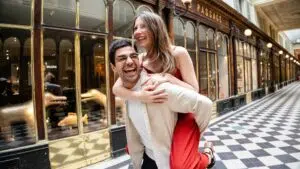 Un homme porte une femme sur son dos dans la galerie couverte Véro Dodat à Paris, au sol carrelé en damier ; tous deux rient et ont l'air heureux.