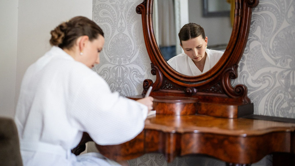 La mariée vêtue d'une robe blanche est assise devant une coiffeuse en bois, en train d'écrire ses vœux pour la cérémonie laïque, sur une feuille de papier, tandis que son reflet se reflète dans un miroir ouvragé.