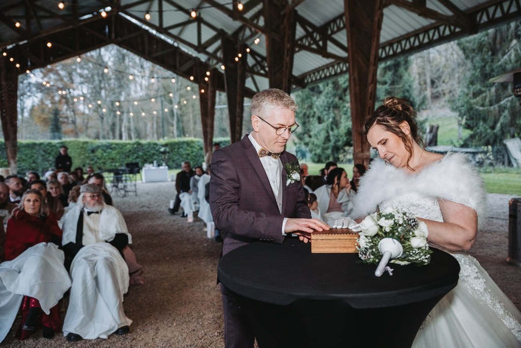 Les mariés se tiennent debout devant une table ronde lors d'une cérémonie de mariage en plein air. Le marié porte un costume ; la mariée porte une étole en fourrure. Les invités sont assis derrière eux, sous des guirlandes lumineuses, et regardent le couple accomplir le rituel de la capsule temporelle.
