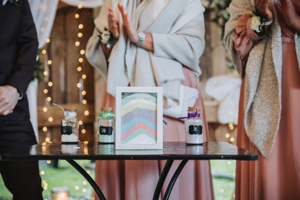 Trois personnes se tiennent derrière une table sur laquelle sont disposés des bocaux remplis de sable coloré et un contenant en verre encadrée contenant des strates de sable de couleur différente. Des guirlandes lumineuses et des drapés décorent l'arrière-plan, créant une ambiance chaleureuse et festive pour la cérémonie laïque.