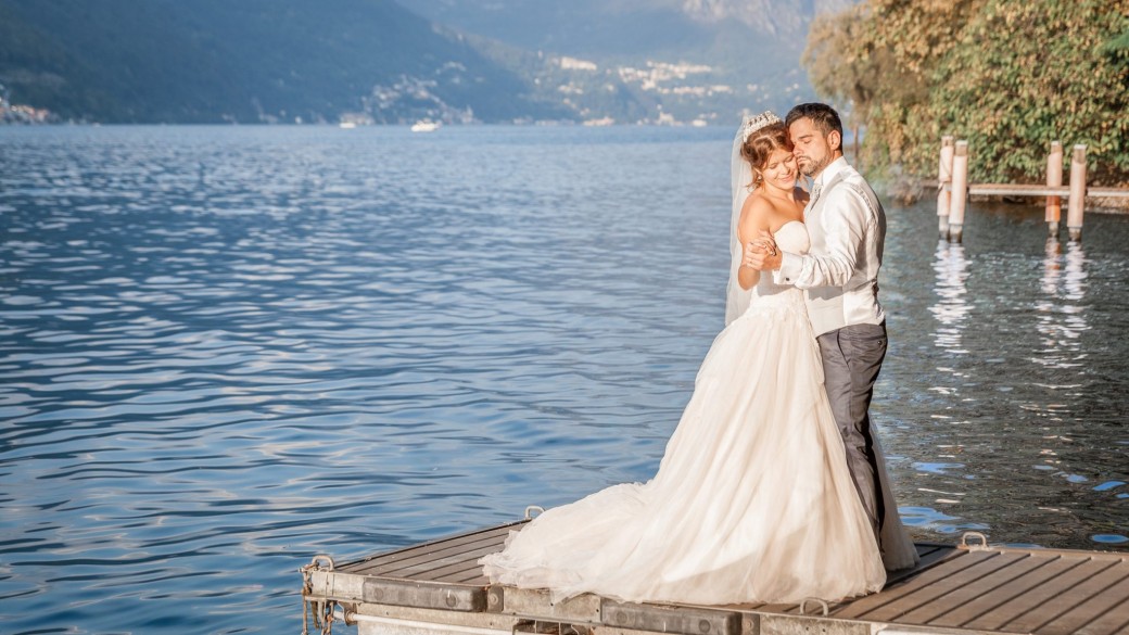 Un couple de mariés s'embrassent sur un quai en bois au bord d'un lac serein, entouré d'une végétation luxuriante et d'un paysage montagneux. La mariée porte une robe blanche fluide et le marié est dans un costume de couleur claire. Le soleil projette une lueur chaleureuse sur la scène.