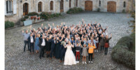 Un grand groupe de personnes, avec un couple de mariés au centre, saluent joyeusement la caméra. Ils sont réunis dans une cour pavée entourée de bâtiments en pierre, pour célébrer un mariage.